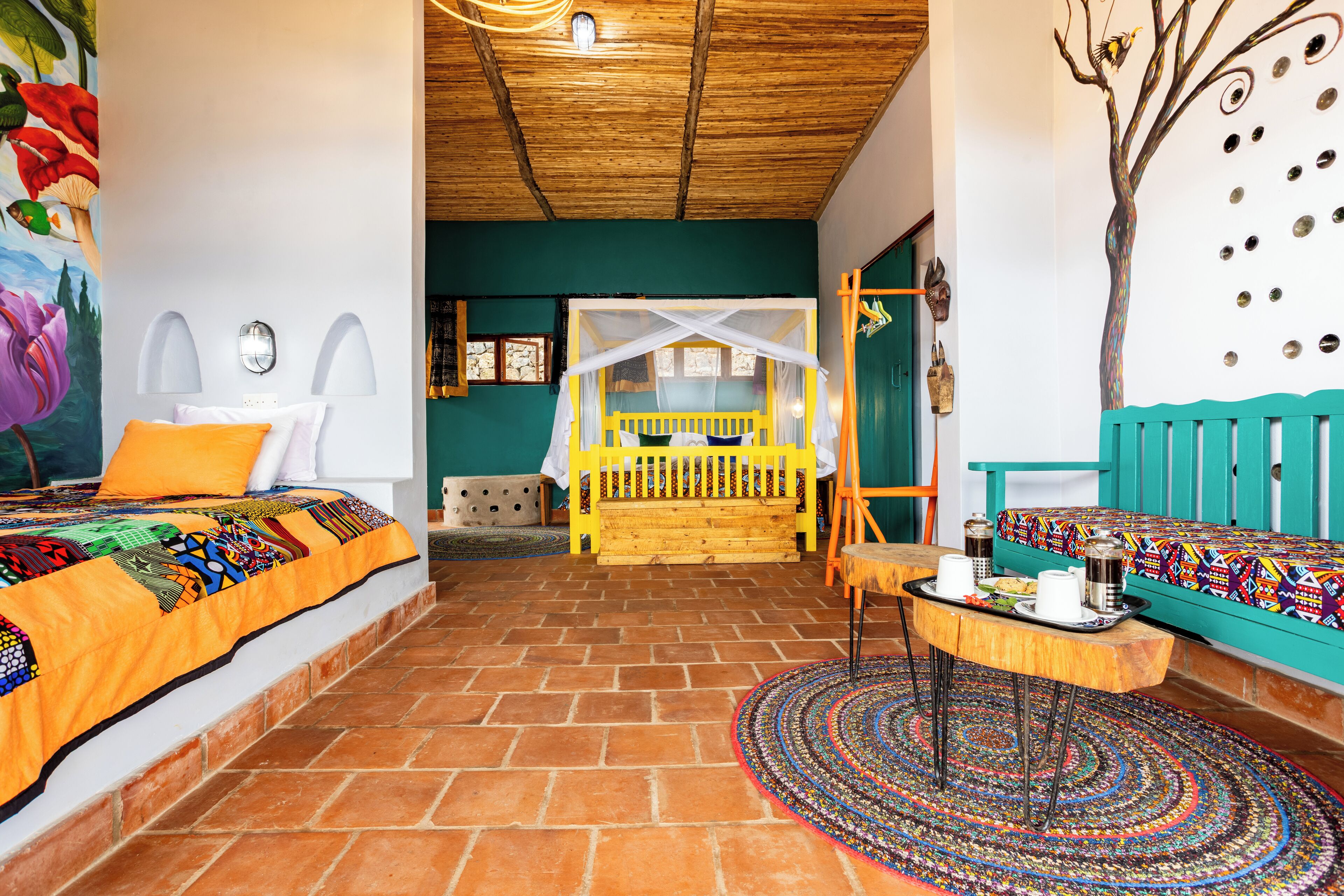 Una habitación con una cama, un banco, una mesa con tazas y una alfombra colorida.