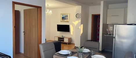 A modern dining area with a round table set for four, a television mounted on the wall, and a kitchen area with a refrigerator.