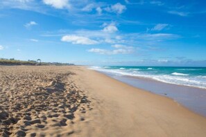 A sandy beach with footprints leading to the water, a clear sky, and a distant structure on the horizon.