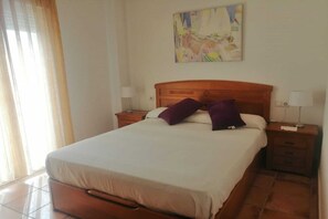 Ein ordentlich bezogenes Bett mit Holzkopfteil, zwei Nachttische mit Lampen und ein farbenfrohes abstraktes Gemälde an der Wand.