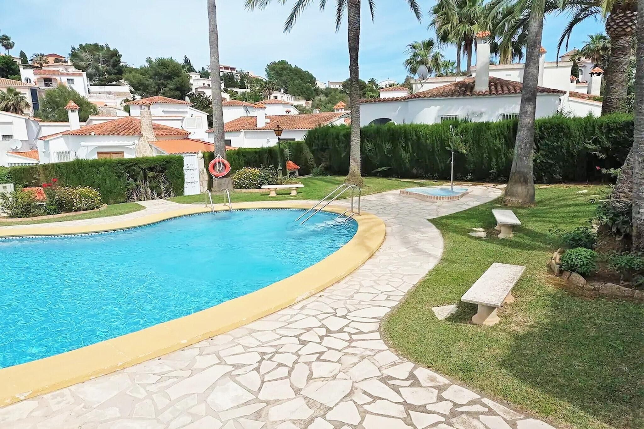 A swimming pool with stone tiles, surrounded by a garden with benches and palm trees.