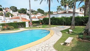 Una piscina con baldosas de piedra, rodeada por un jardín con bancos y palmeras.