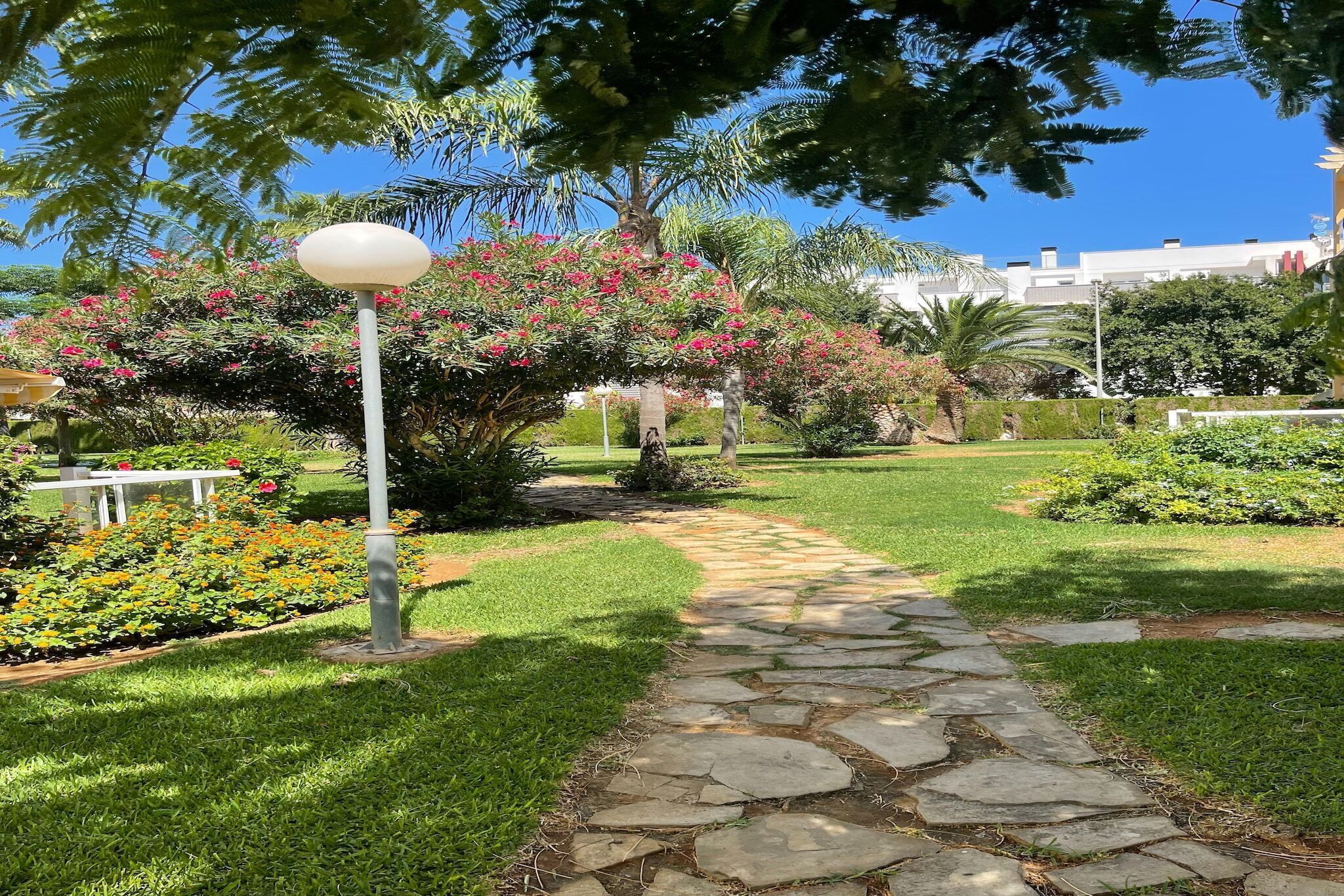 A stone pathway leading through a well-maintained garden with various plants and a lamp post.