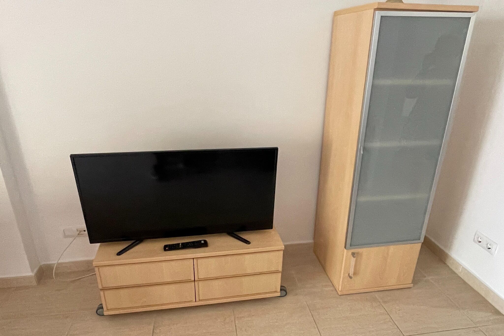 A flat-screen TV on a wooden TV stand with a remote control, next to a tall, narrow cabinet with a glass door.