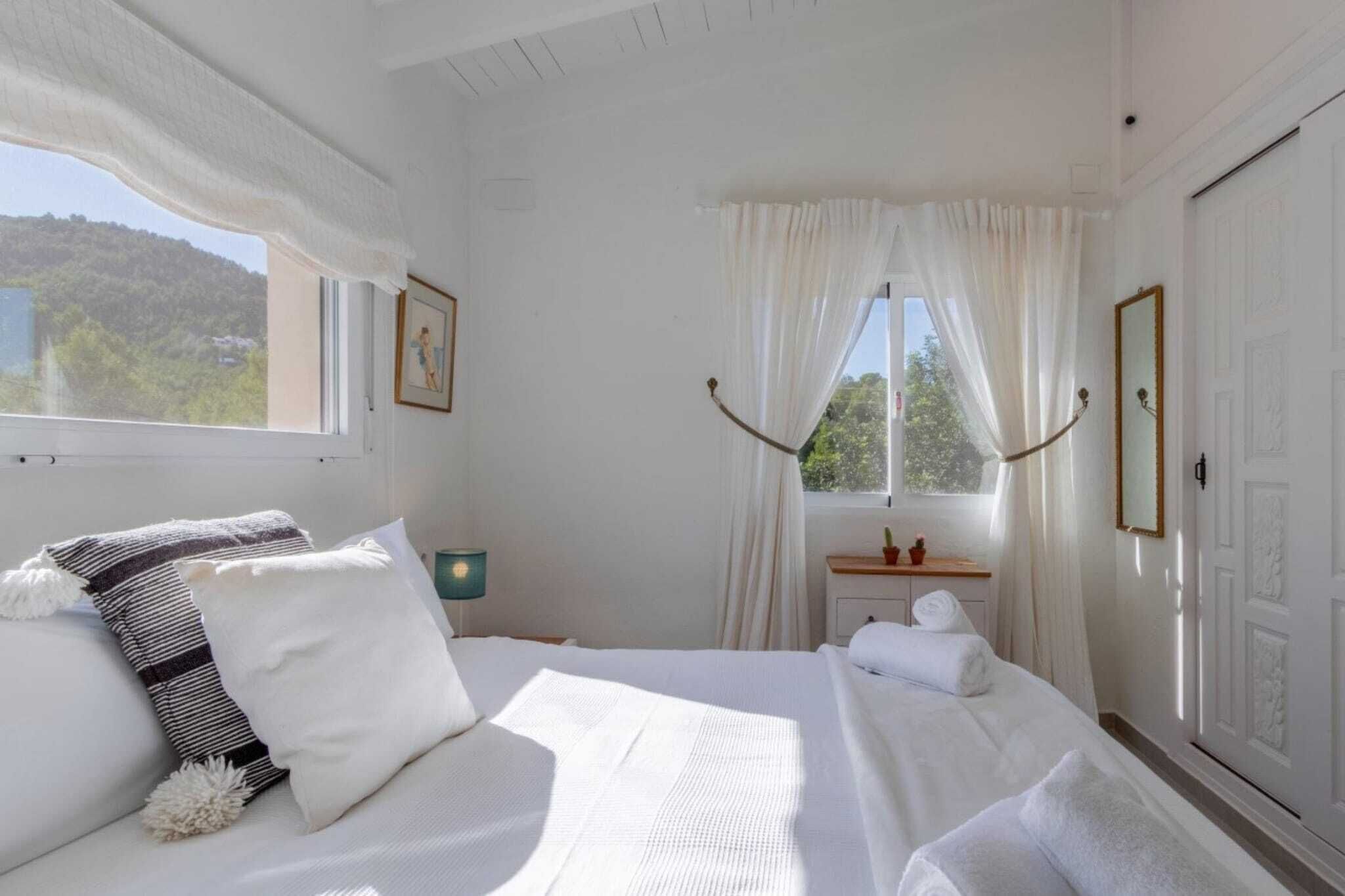 Ein Schlafzimmer mit einem großen Bett, einem Nachttisch mit Lampe, einem Fenster mit Vorhängen und Blick ins Grüne.