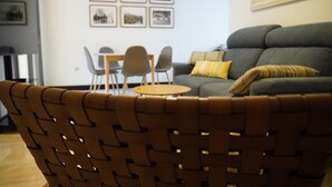 A living room with a brown woven chair, a grey sofa, and a small wooden table. There are framed pictures on the wall and a patterned tapestry above the sofa.