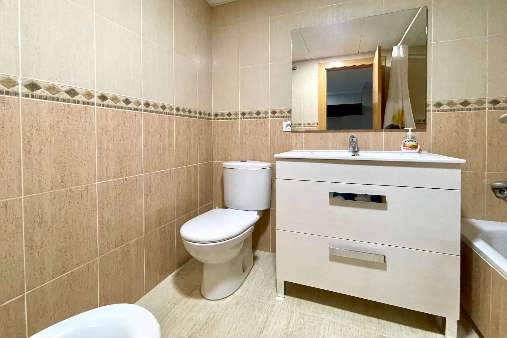 Une salle de bain avec des murs carrelés de couleur beige, des toilettes blanches, un meuble-lavabo blanc avec un miroir et une baignoire blanche.