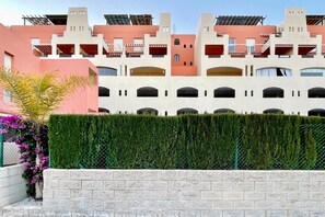 A multi-story building with balconies and a green hedge in the foreground.