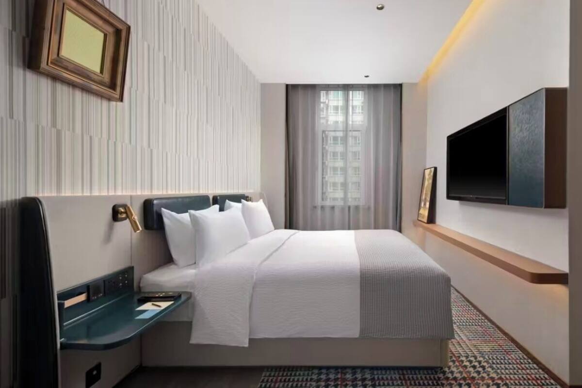 A modern hotel room with a large bed, a bedside table, a TV mounted on the wall, and a window with curtains.
