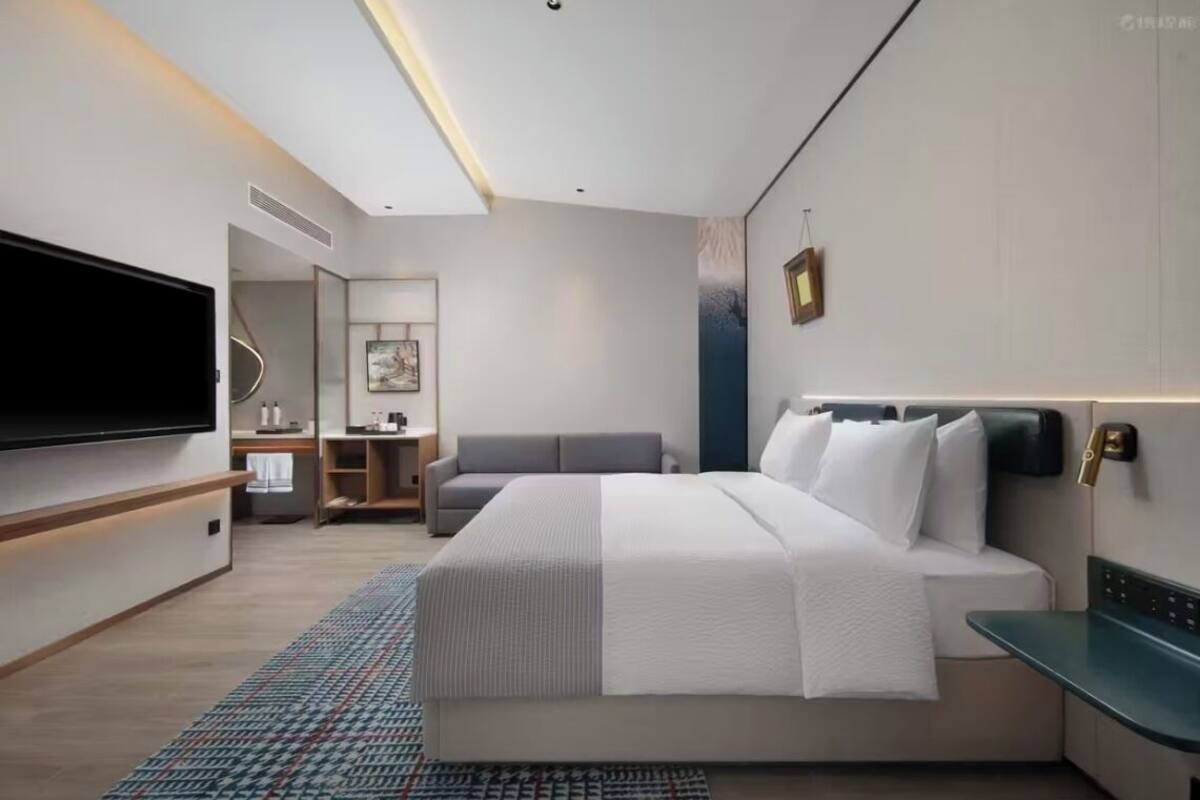 A modern hotel room with a large bed, bedside tables, a flat-screen TV, a sofa, and a small kitchenette.