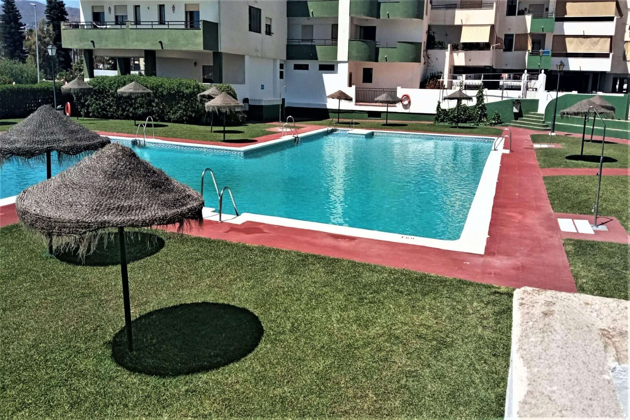 Une piscine bordée de rouge, entourée de pelouse et ombragée par des parasols en chaume.