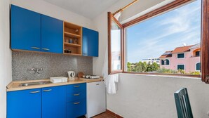 A compact kitchen with blue cabinets, a small white refrigerator, and a window overlooking a residential area. - Apartment Šibenik near Beach Gaj (Šibenik-Brodarica)