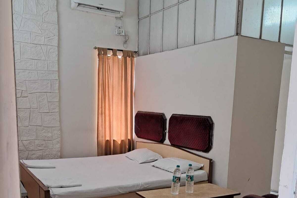 Quarto de hotel compacto com cama, uma mesinha com garrafas de água, um sofá e um aparelho de ar condicionado na parede.