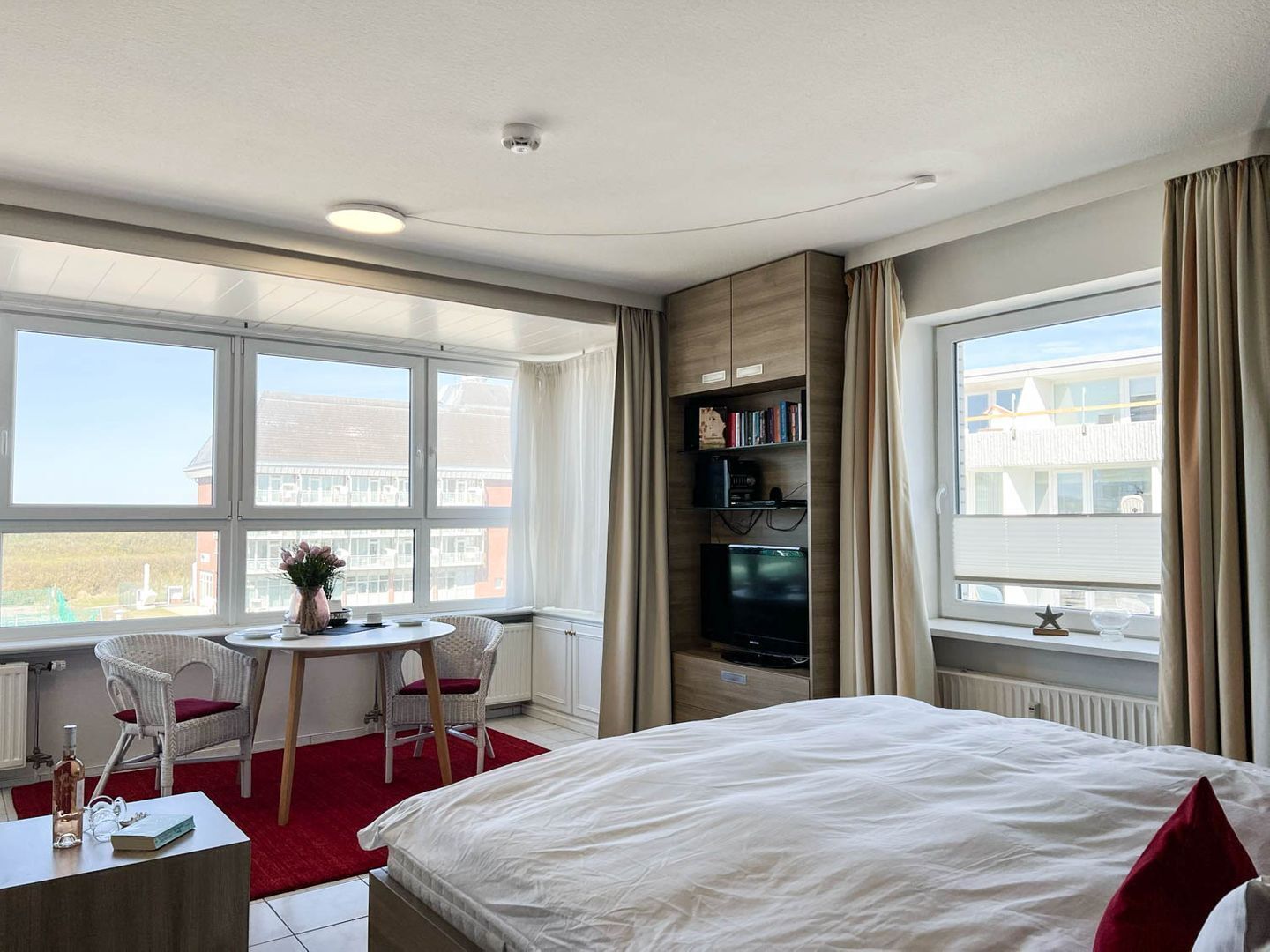 A hotel room with a bed, a desk with a chair, a small table, a bookshelf, and large windows offering a view of buildings outside.