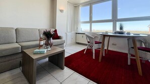 Un salon moderne avec un canapé gris, une table basse en bois et un tapis rouge. On y trouve également un coin repas avec des chaises et une table en osier. De grandes fenêtres offrent une vue sur l’extérieur.