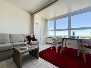 A modern living room with a grey sofa, a wooden coffee table, and a red rug. There is a dining area with wicker chairs and a table. Large windows provide a view of the outdoors.