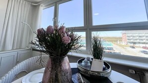 Une pièce avec une grande fenêtre, un vase rempli de fleurs, un plateau avec des pots et une vue sur un immeuble et un parking à l’extérieur.