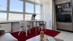 Un salon moderne avec une grande fenêtre, un coin repas avec des chaises en osier, une moquette rouge, un téléviseur et une bibliothèque.