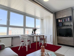 A modern living room with a large window, a dining area with wicker chairs, a red carpet, a television, and a bookshelf.