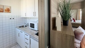Une cuisine moderne avec des meubles blancs, un four à micro-ondes et un évier. On aperçoit une plante sur une étagère en bois et un lit dans la pièce adjacente.