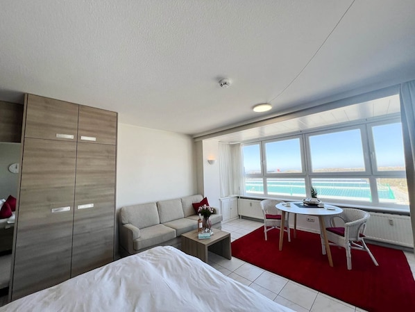 A modern hotel room with a bed, a sofa, a dining table with chairs, a red carpet, and a large window offering a view of the sea.