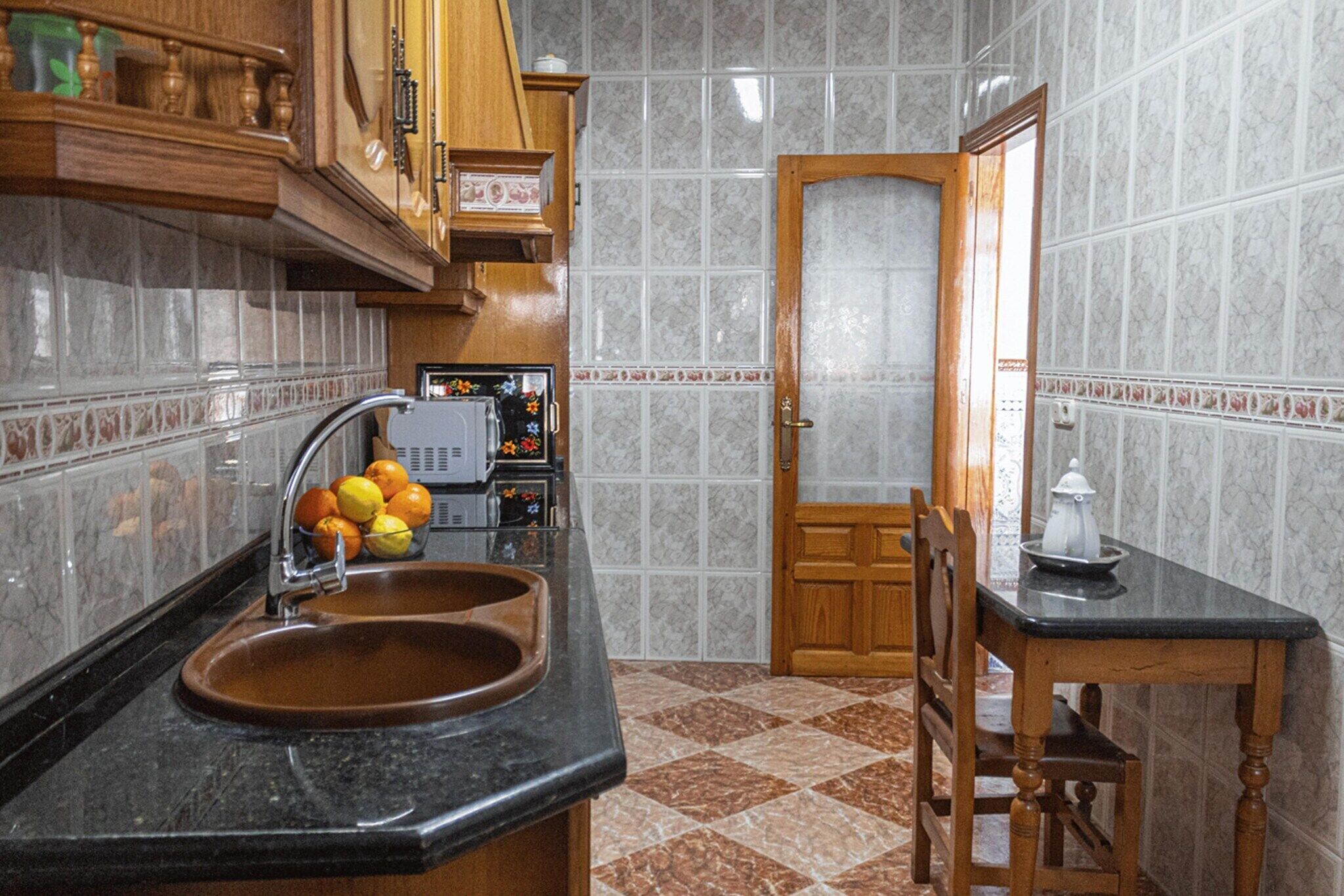 Une cuisine dotée d’un plan de travail en granit, d’un évier, d’un meuble en bois, d’une petite table avec une chaise, et d’une porte donnant sur une autre pièce.
