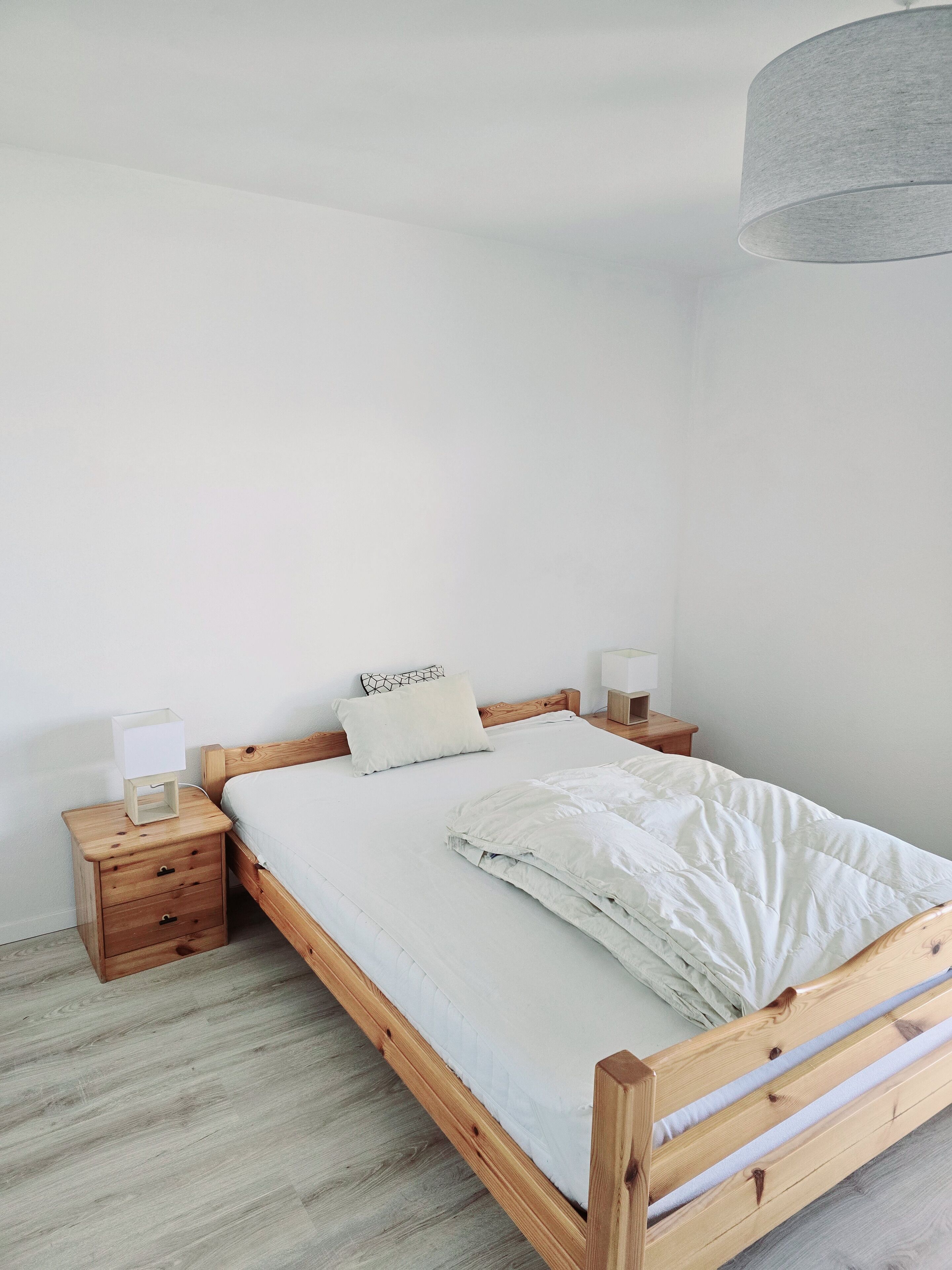 Ein schlicht eingerichtetes Schlafzimmer mit einem hölzernen Bett, weißer Bettwäsche, einem Nachttisch mit Lampe und einer Pendelleuchte.