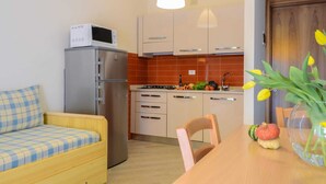 A compact kitchen with a refrigerator, microwave, and stove. There's a wooden dining table with chairs and a small sofa with a plaid cover. A vase with flowers is on the table. - Apartment for 5 guests in Pietra Ligure (Pietra Ligure)