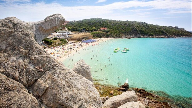 A coastal beach with clear turquoise waters, rocky cliffs, and a densely populated beach area with umbrellas and people. - Apartment in Olbia with Private Terrace (Olbia)