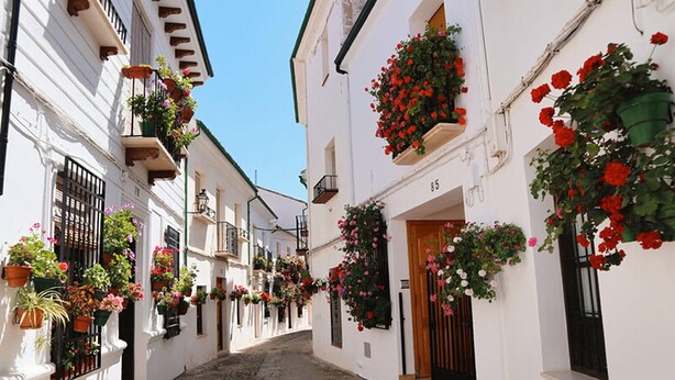 A narrow cobblestone street lined with white buildings, each adorned with flower boxes and balconies.