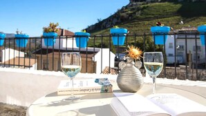 A table with two glasses of white wine, a book, and a vase with a decorative item, set against a scenic view of a hill and buildings.