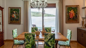 A dining area with a wooden table and chairs, two paintings, a chandelier, and a view of the sea through large windows. - Holiday home in Córdoba with Swimming pool (Zambra)