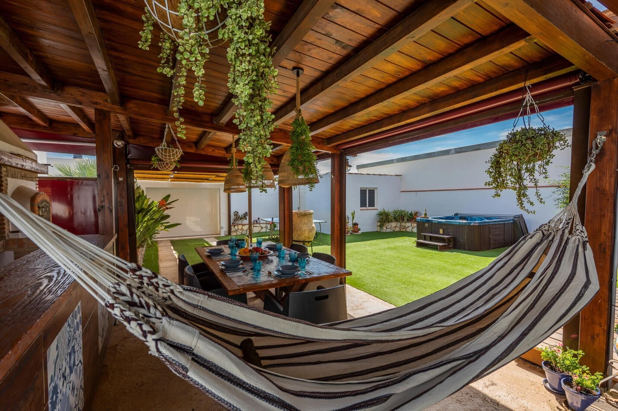 A wooden outdoor patio with a hammock, hanging plants, a table set for dining, and a hot tub in the background.