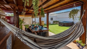 A wooden outdoor patio with a hammock, hanging plants, a table set for dining, and a hot tub in the background. - Holiday home in Córdoba with Swimming pool (Zambra)