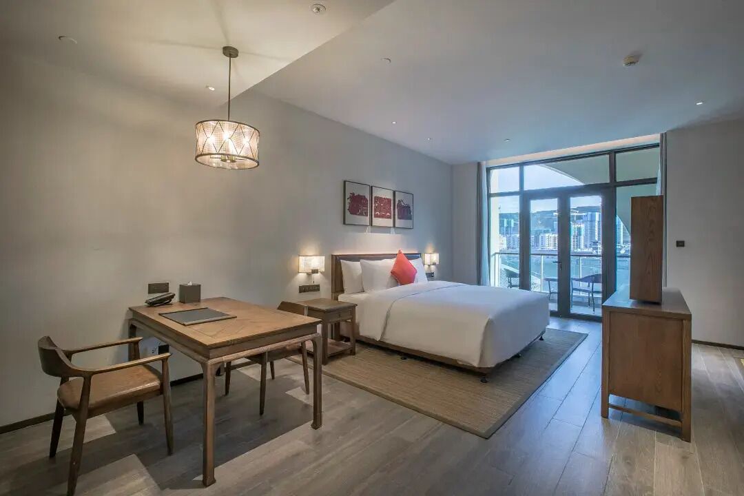 Een moderne hotelkamer met een groot bed, een eettafel, stoelen, een bureau en uitzicht op de stad door grote ramen.