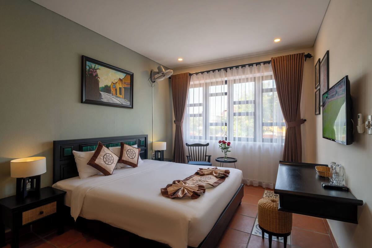 A hotel room with a large bed, a desk, a chair, a TV, and a window with curtains.