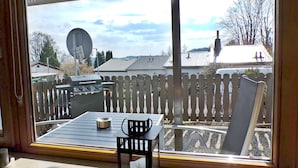 Blick durch ein Fenster in einen Hinterhof mit Holzzaun, Grill und Tisch mit Stühlen.