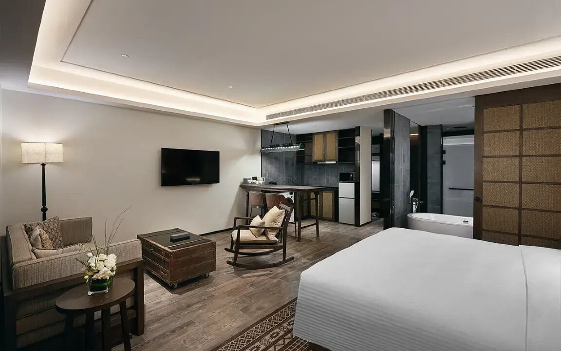 A modern hotel room with a large bed, a sofa, a small table, a rocking chair, a TV, and a kitchenette area.