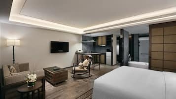 A modern hotel room with a large bed, a sofa, a small table, a rocking chair, a TV, and a kitchenette area.