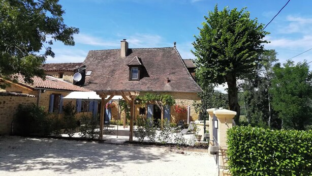 Una casa de piedra tradicional con techo de tejas, un porche cubierto y un jardín con diversas plantas y un pequeño árbol.
