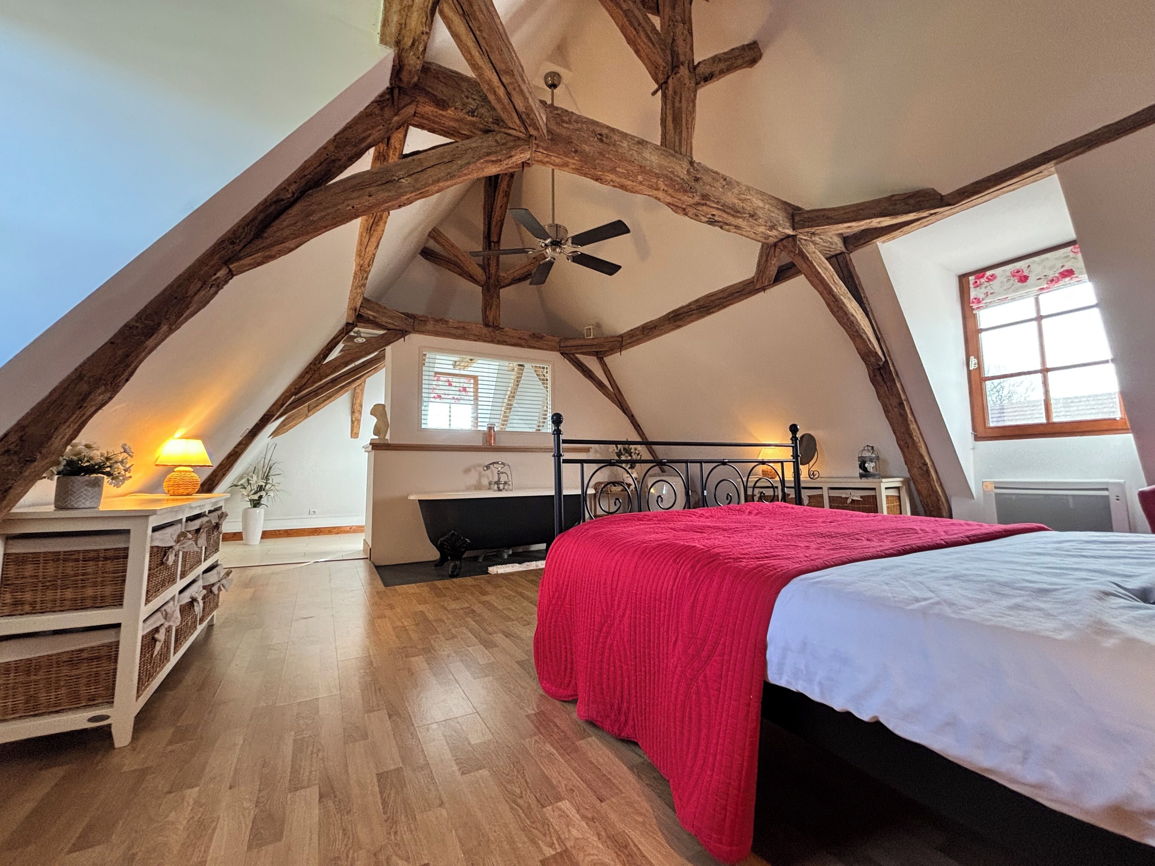 A bedroom with a sloped ceiling, exposed wooden beams, a bed with a red blanket, a fireplace, and a window with blinds.