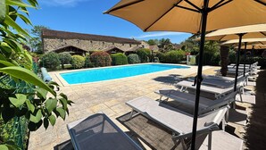 Una piscina con sillas de descanso y sombrillas, rodeada de edificios de piedra y vegetación.