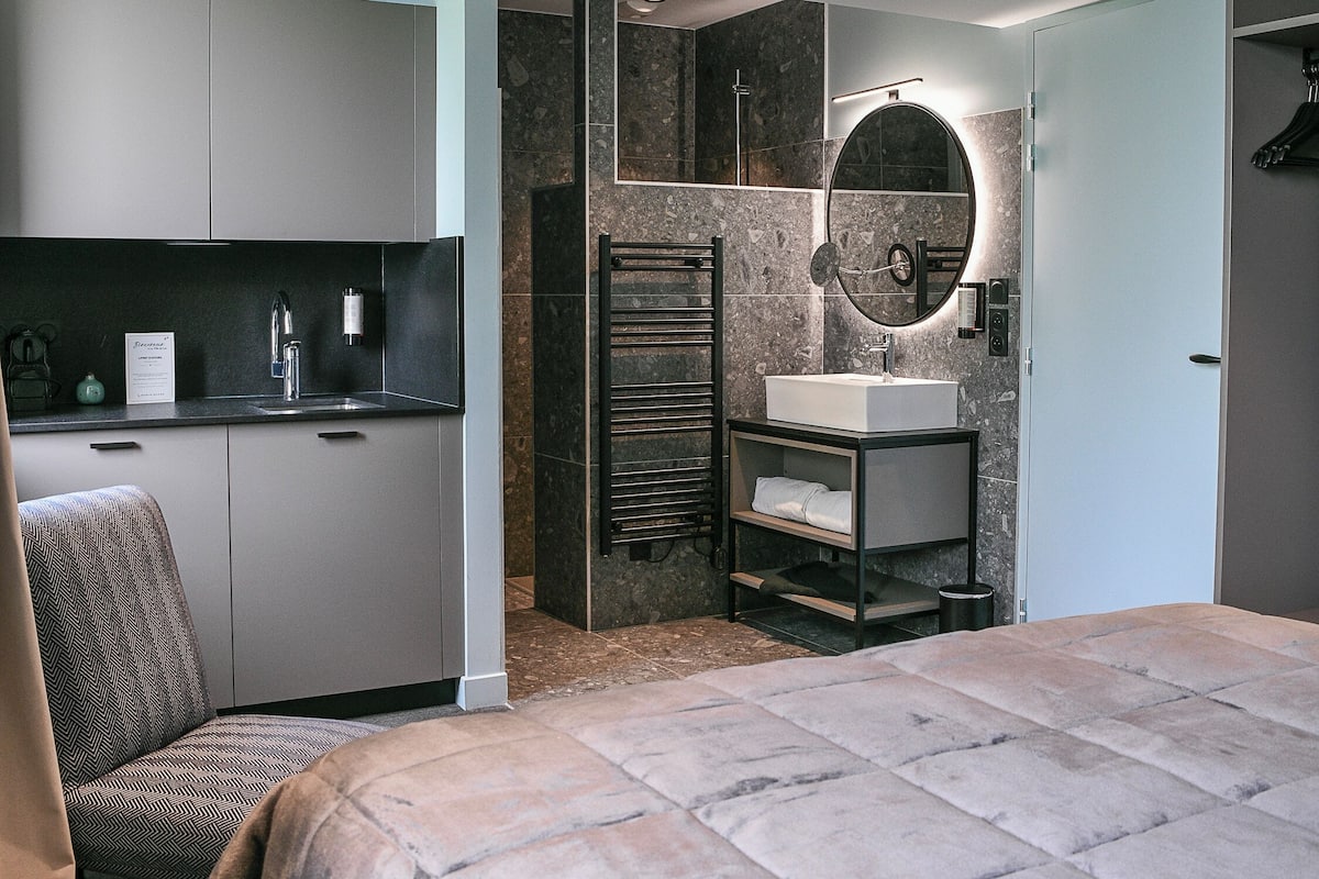 Une chambre moderne dotée d’un grand lit, d’une armoire intégrée, d’une salle de bain avec un lavabo et un miroir, et d’un coin salon avec un fauteuil.