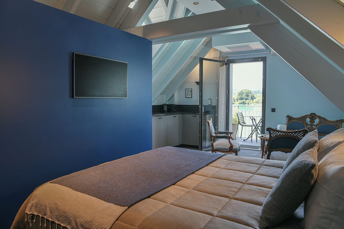 Une chambre avec un grand lit, un mur bleu et une vue sur un balcon aménagé avec du mobilier d’extérieur.