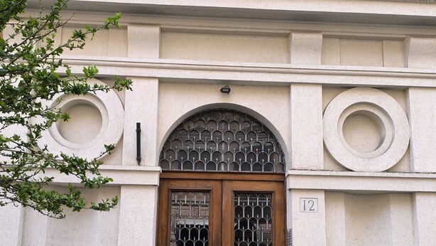 A double wooden door with a metal grille, a small circular window above, and a number "12" on the wall to the right.
