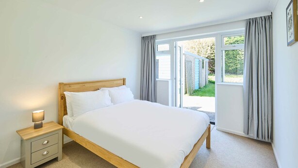 A bedroom with a wooden bed, a nightstand with drawers, a bedside lamp, and a large window with curtains. - Host Stay Park View (Thetford)