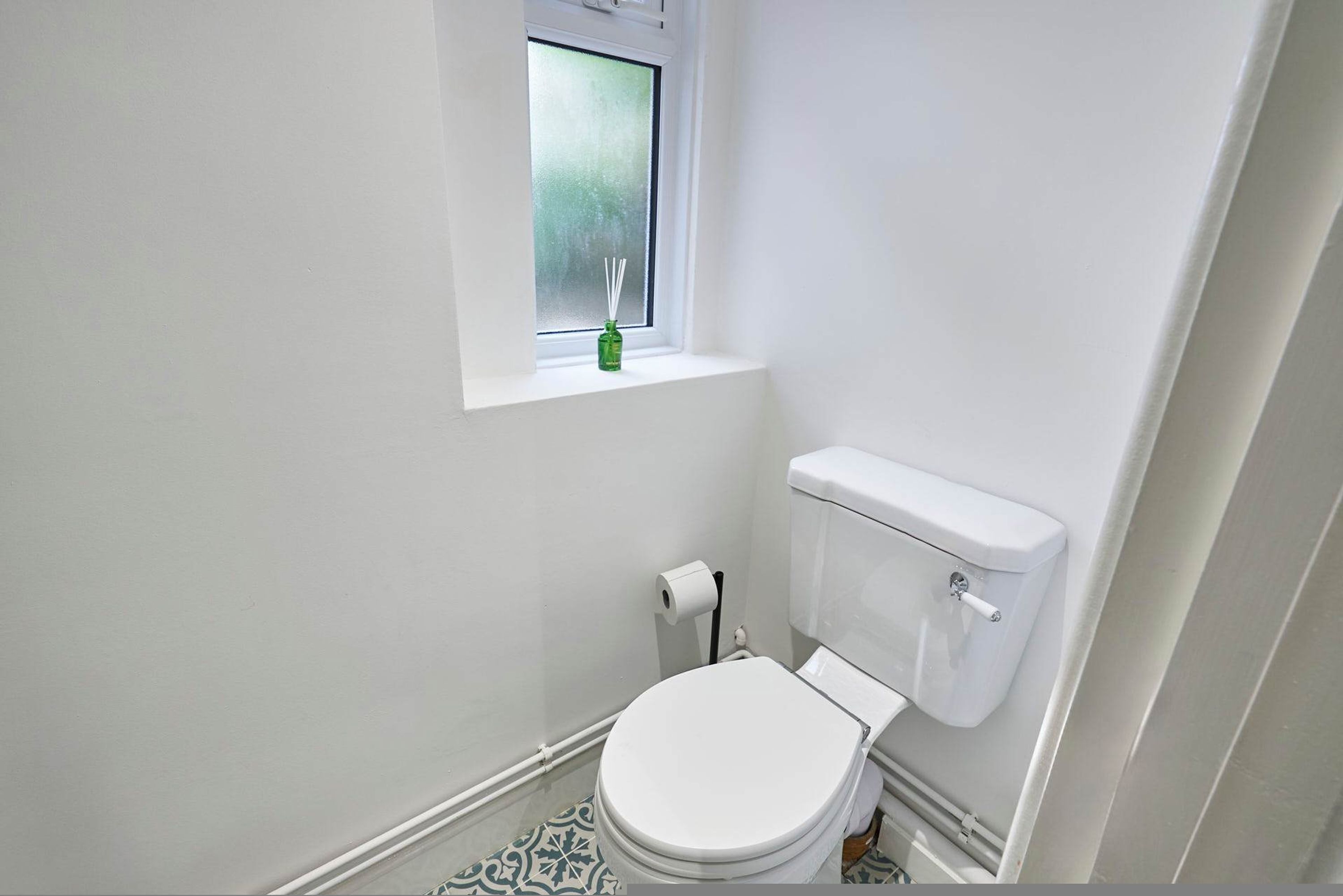 Une petite salle de bain blanche, avec des toilettes, une fenêtre ornée d’un vase vert et un carrelage au sol à motifs.