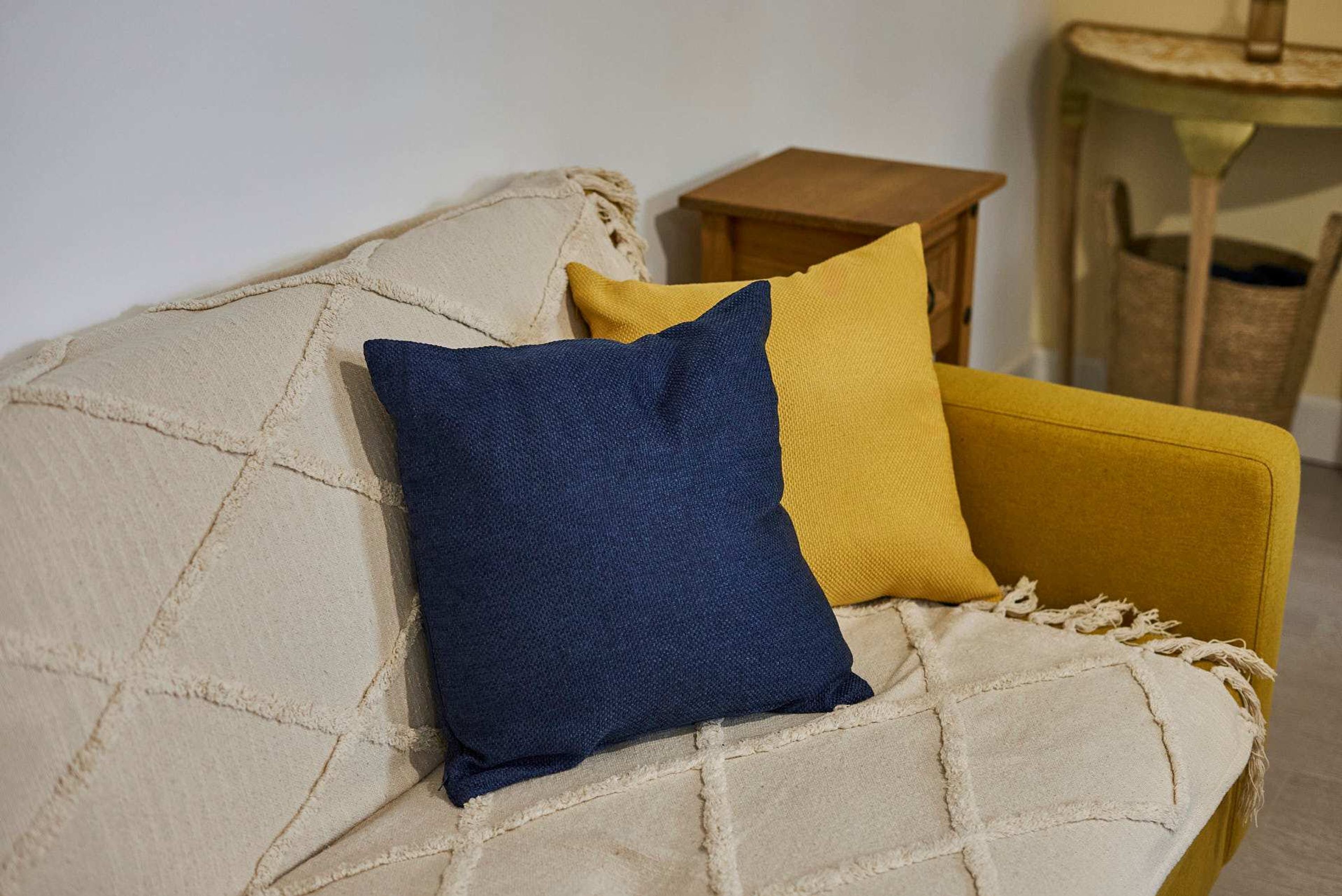 En beige soffa med ett quiltat mönster och två kuddar, en blå och en gul, i ett rum med ett litet sidobord i trä och en stol av rotting.