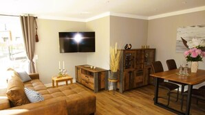 Un salon avec un canapé marron, une table à manger en bois avec des chaises, un téléviseur fixé au mur et un meuble contenant des objets décoratifs.