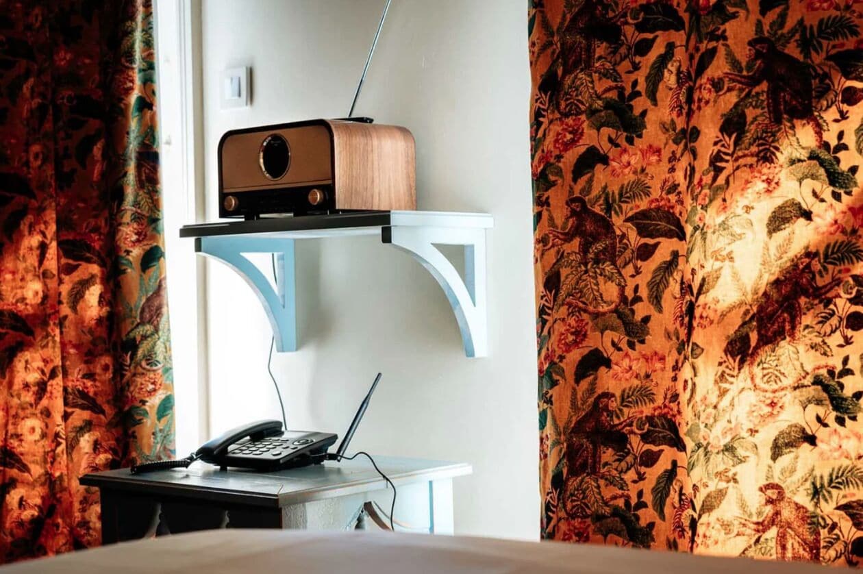 A room with a vintage radio on a blue shelf, a desk with a laptop and a telephone, and floral-patterned curtains.